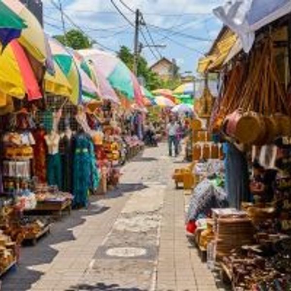 Voyage en Asie — Marché d'Art d'Ubud à Bali : allée animée bordée de stands colorés avec vêtements, sacs en paille, objets en bois sculpté et parasols tropicaux.