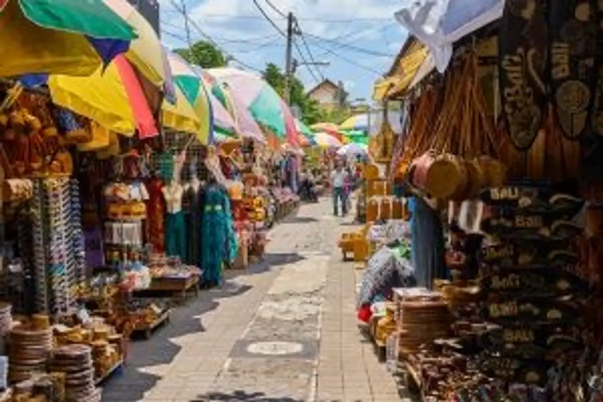 Voyage en Asie — Marché d'Art d'Ubud à Bali : allée animée bordée de stands colorés avec vêtements, sacs en paille, objets en bois sculpté et parasols tropicaux.