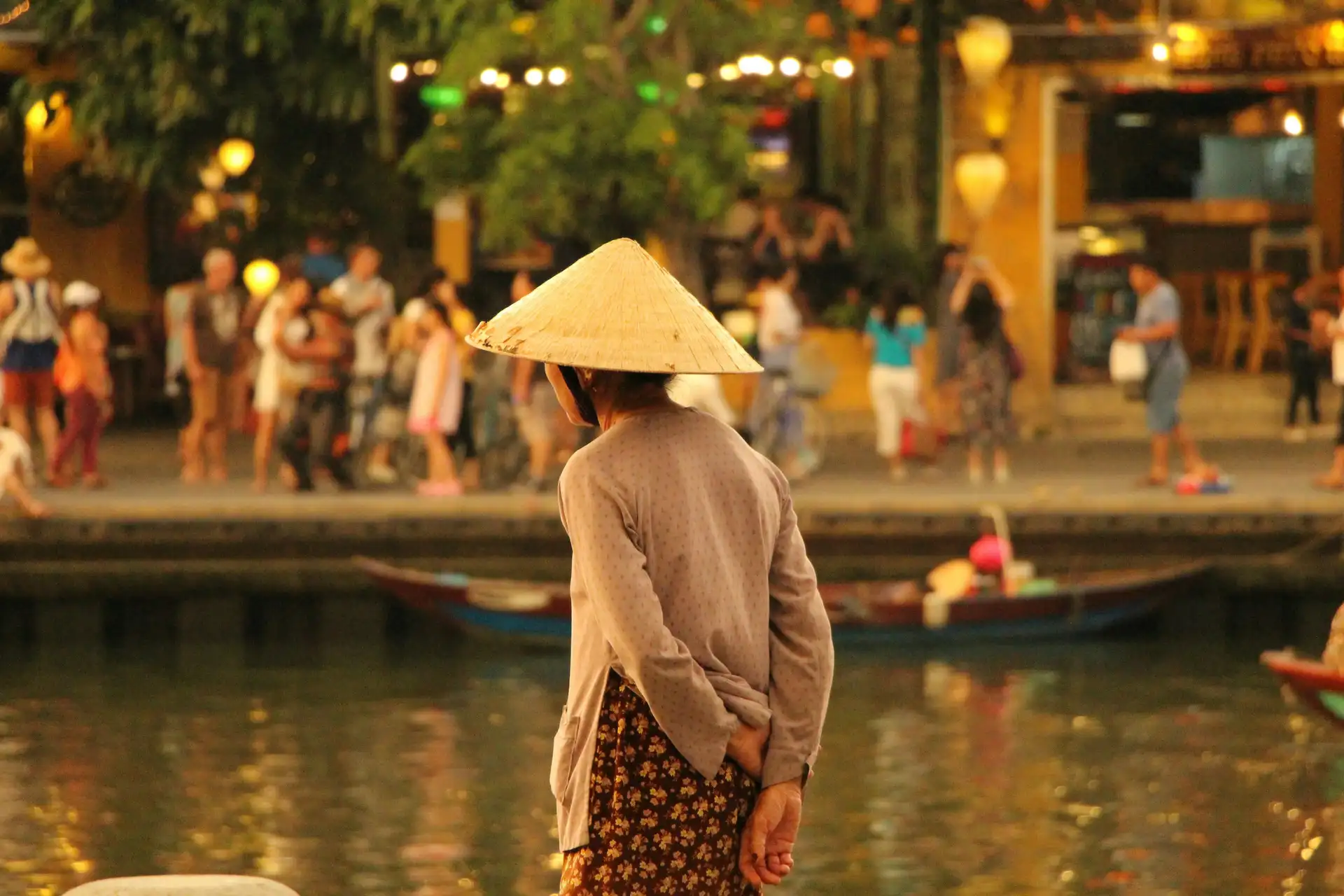 Voyage au Vietnam – Ambiance nocturne sereine au bord de la rivière à Hoi An avec des lanternes illuminées et une femme portant un chapeau conique traditionnel.