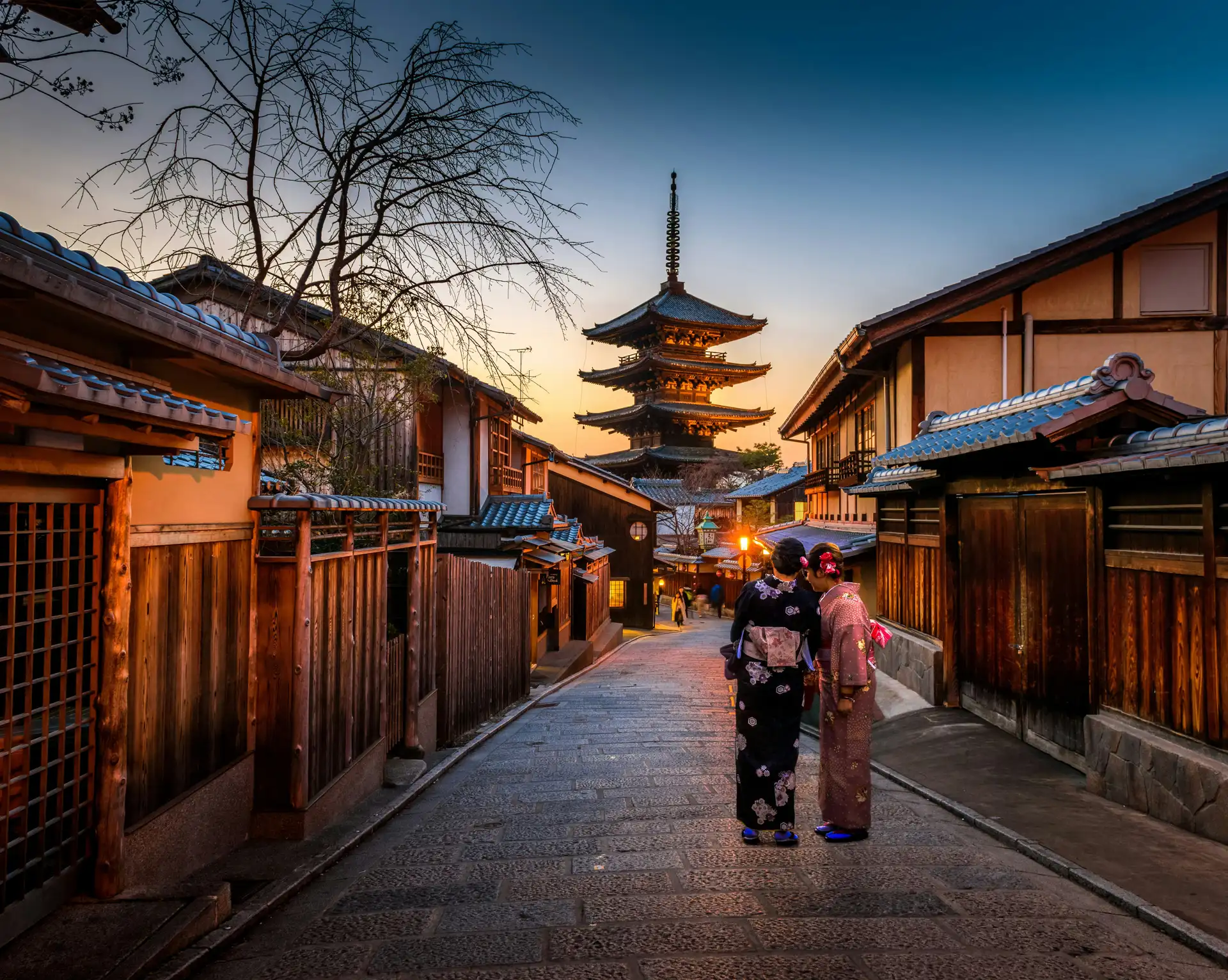 Rue traditionnelle du quartier d'Higashiyama à Kyoto bordée de maisons en bois avec la pagode Yasaka en arrière-plan au coucher du soleil.