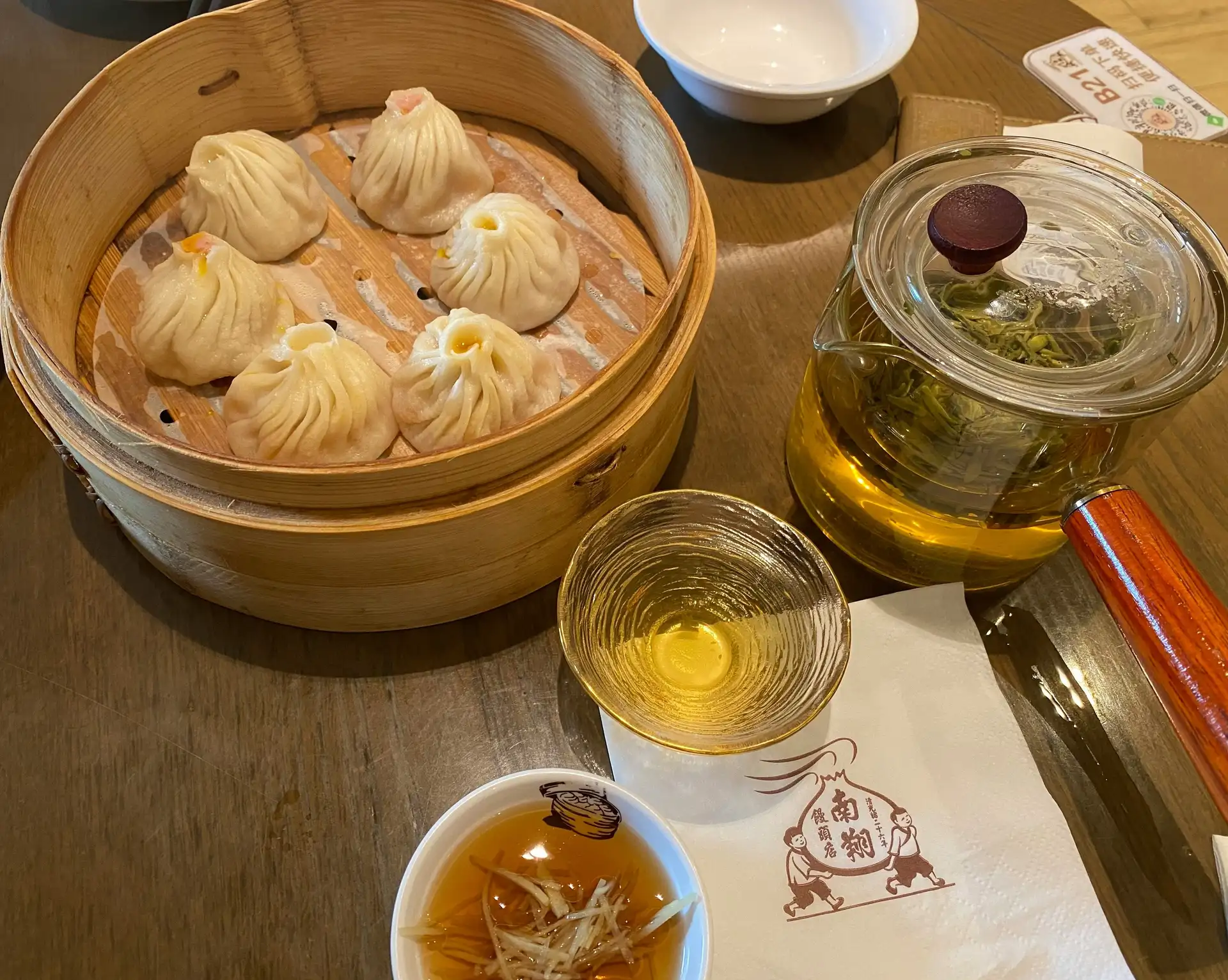 Un panier vapeur en bambou contenant plusieurs raviolis chinois Xiaolongbao chauds, posé sur une table en bois à côté d'une théière transparente et d'une tasse de thé.