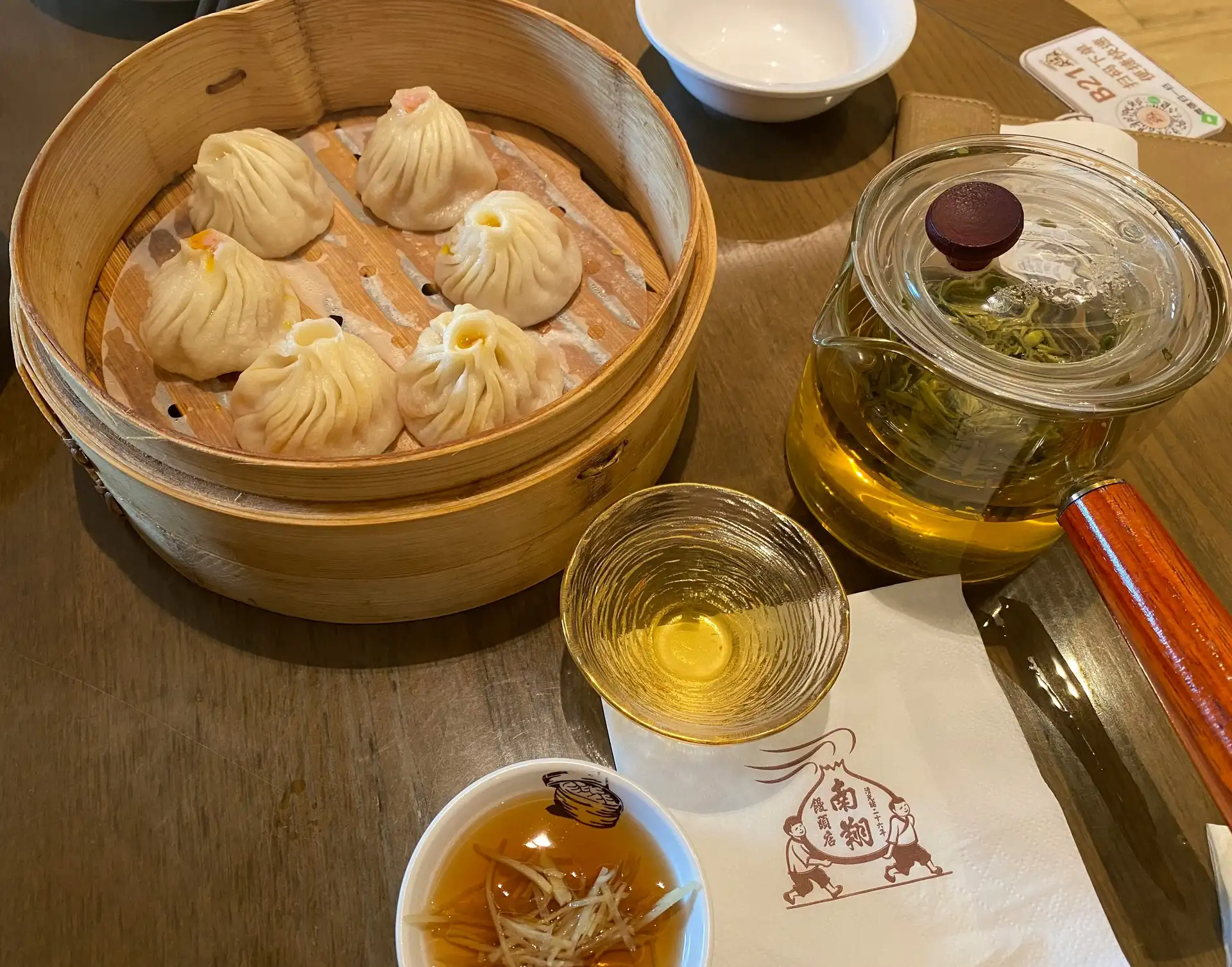 Un panier vapeur en bambou contenant plusieurs raviolis chinois Xiaolongbao chauds, posé sur une table en bois à côté d'une théière transparente et d'une tasse de thé.