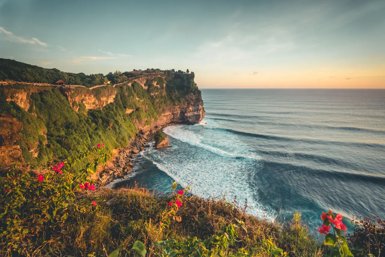 Une haute falaise rocheuse escarpée et verdoyante, typique d'Uluwatu, plongeant dans l'océan Indien aux vagues régulières sous un ciel de fin de journée à Bali.