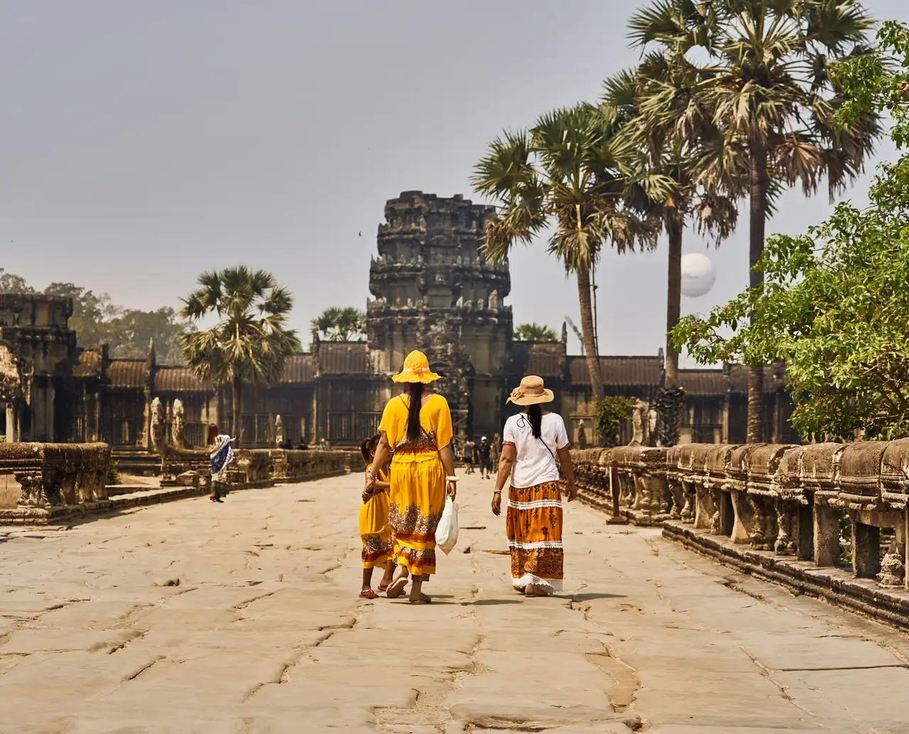 Vue de dos de deux voyageurs explorant le site archéologique d'Angkor Wat en marchant vers les structures monumentales en pierre grise entourées de palmiers au Cambodge.