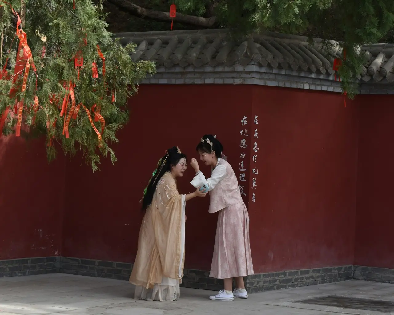 Deux jeunes femmes en vêtements traditionnels chinois partagent un moment de complicité et rient ensemble devant un grand mur rouge historique en Chine.