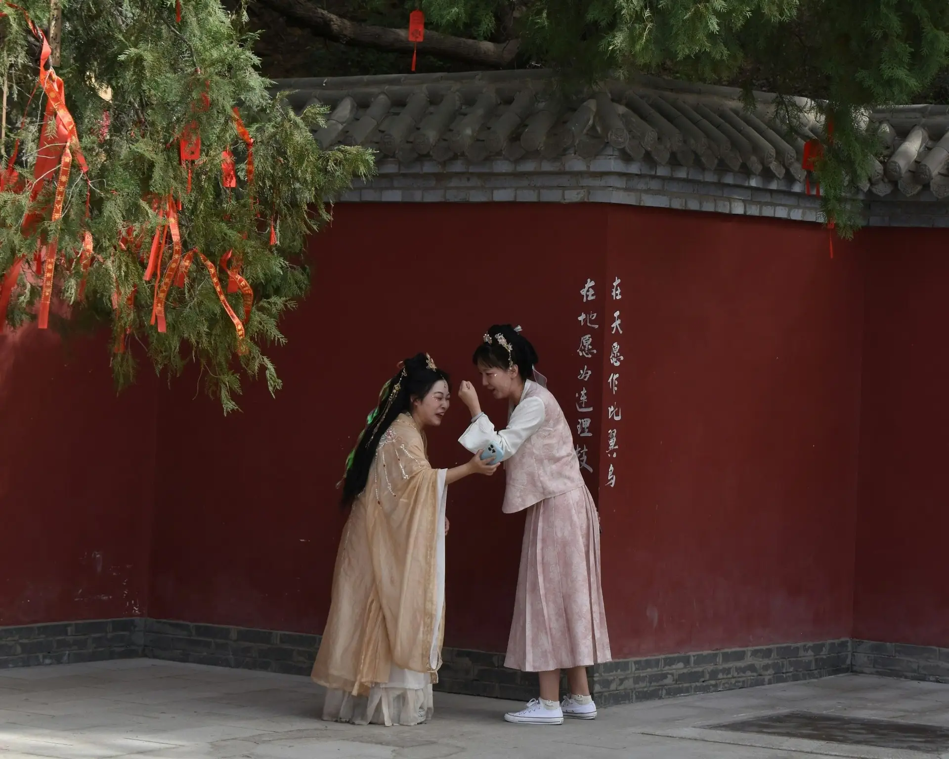 Deux jeunes femmes en vêtements traditionnels chinois partagent un moment de complicité et rient ensemble devant un grand mur rouge historique en Chine.