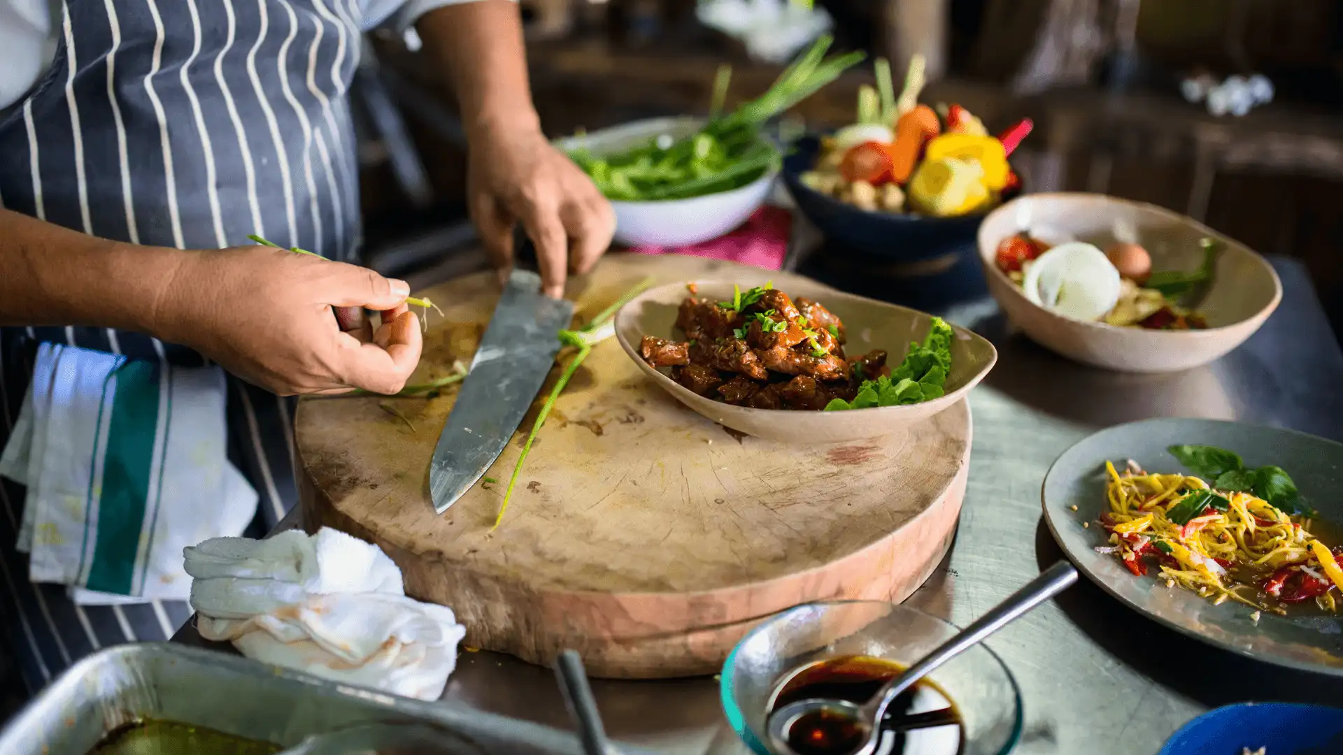 chef cuisinier en tablier raye decoupant des herbes fraiches et legumes locaux sur une grande planche en bois lors d un cours de cuisine traditionnelle au cambodge