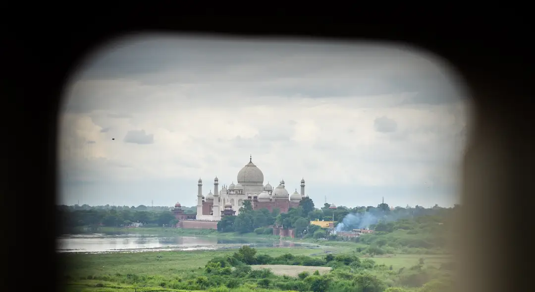 Une vue lointaine du Taj Mahal entouré de végétation et bordé par la rivière Yamuna, cadrée à travers l'ouverture en pierre d'une fenêtre fortifiée du Fort Rouge d'Agra sous un ciel nuageux.