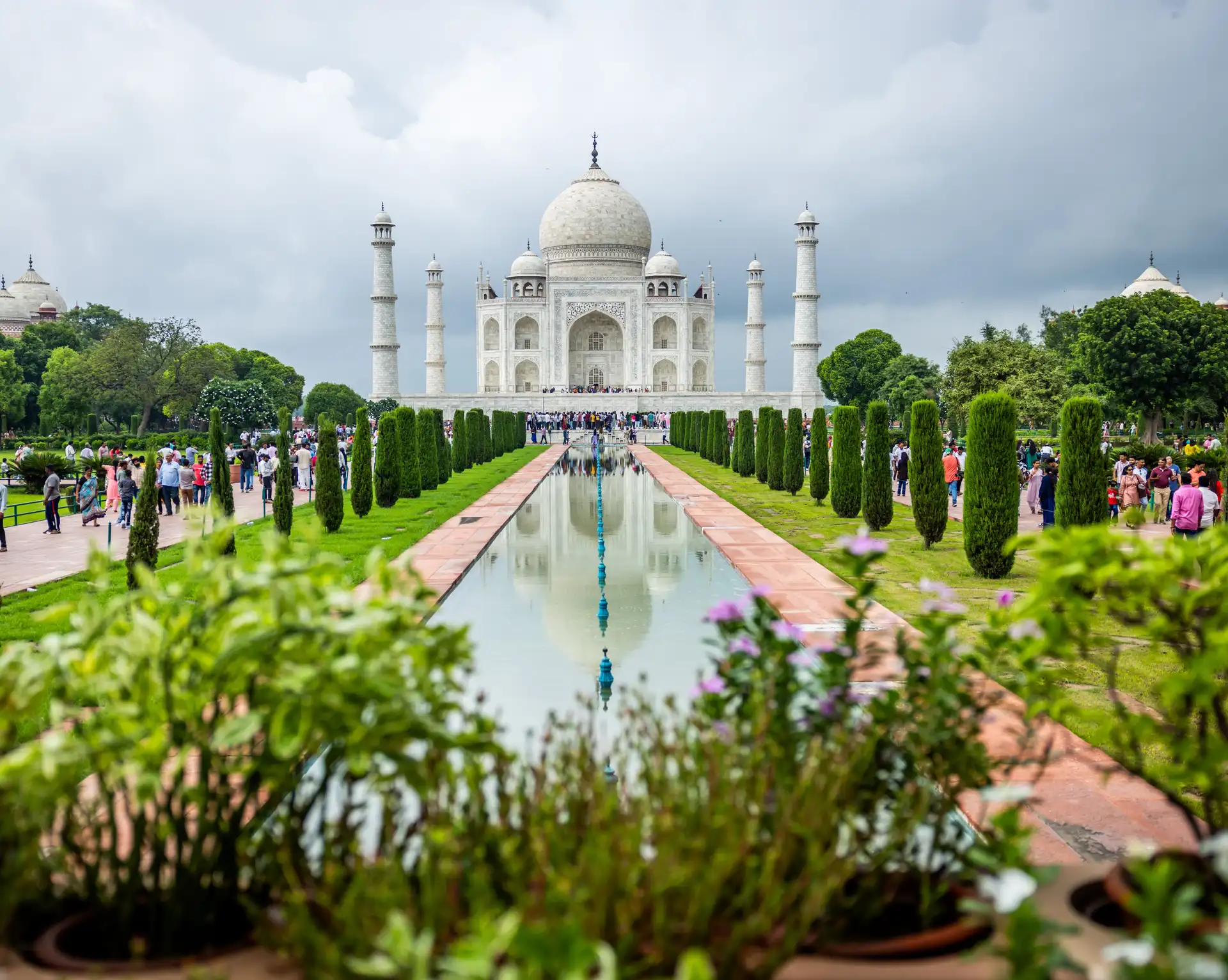 La façade symétrique en marbre blanc du Taj Mahal à Agra se reflétant sur le canal central au milieu des jardins arborés, sous un ciel nuageux en Inde.