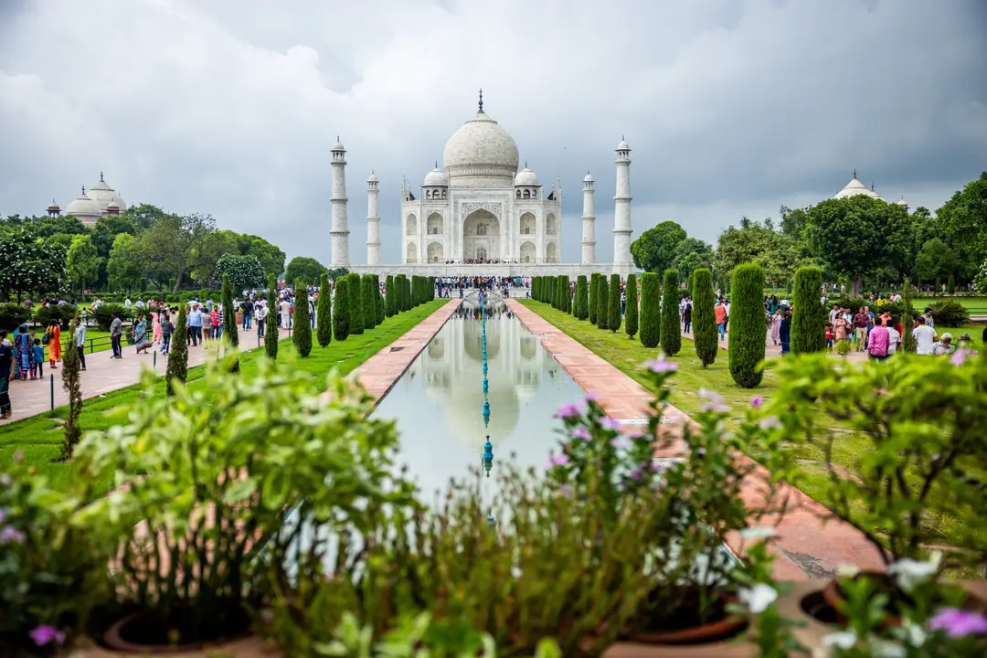La façade symétrique en marbre blanc se reflétant sur le canal central au milieu des jardins arborés, sous un ciel nuageux en Inde.