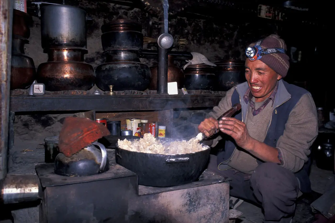 Hôte ladakhi souriant cuisinant des momos traditionnels dans une cuisine typique du Ladakh.