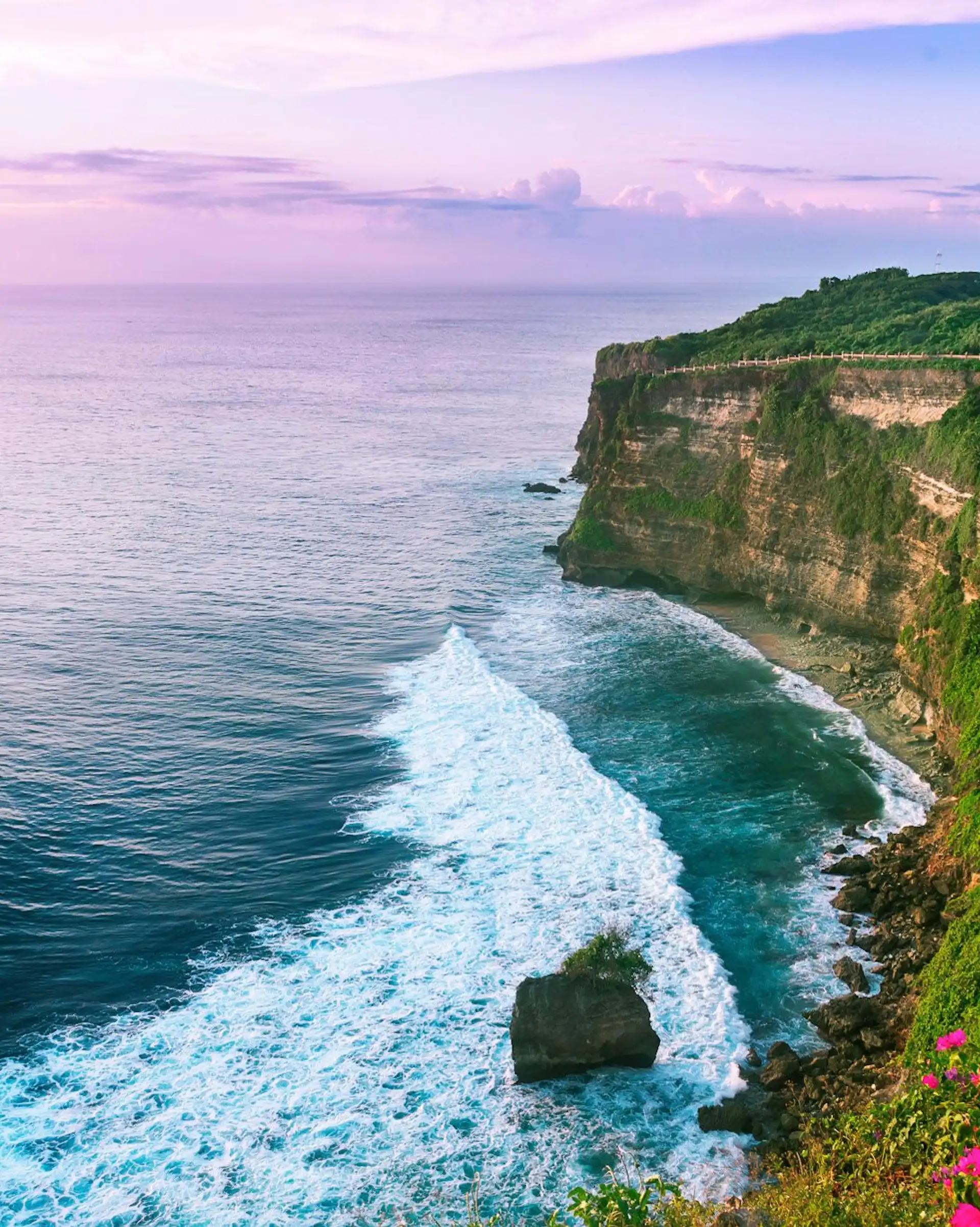 Une vue panoramique des hautes falaises verdoyantes d'Uluwatu plongeant dans l'océan Indien avec des vagues blanches, sous un ciel de fin de journée à Bali.