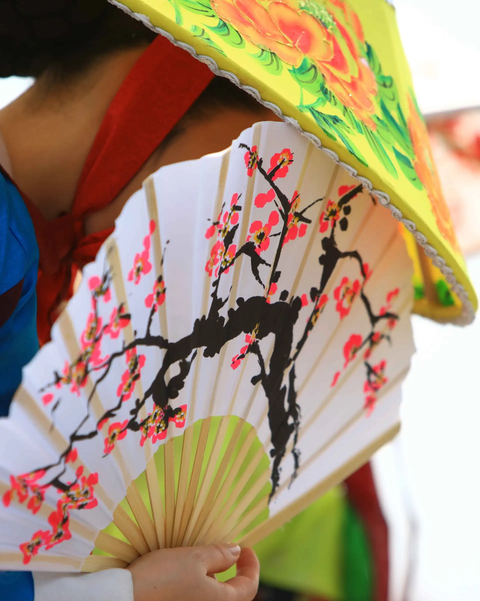 Une personne en tenue traditionnelle de profil portant un grand chapeau conique fleuri et tenant un éventail peint de cerisiers en fleurs en Corée du Sud.