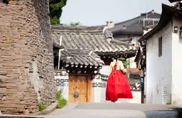 Une personne en hanbok traditionnel marchant dans une ruelle pittoresque bordée de maisons traditionnelles coréennes (hanoks) à Jeonju.