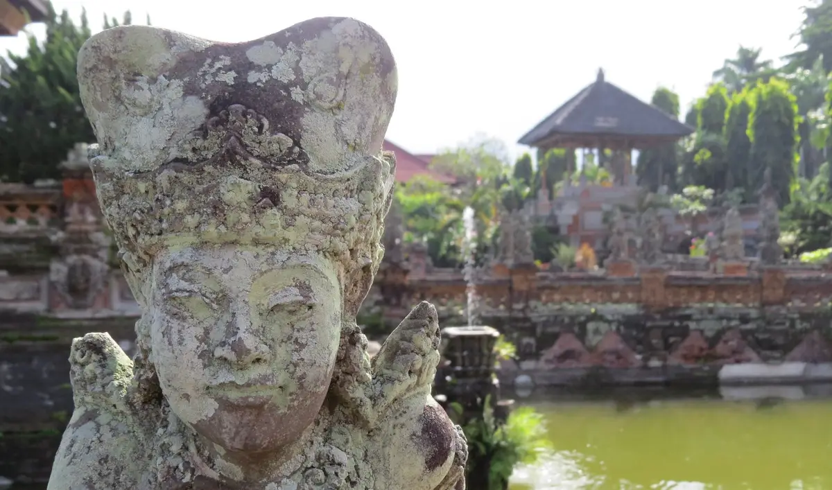 Gros plan sur une statue en pierre sculptée traditionnelle balinaise érodée par le temps, avec en arrière-plan un pavillon historique et les jardins d'un palais royal à Bali.