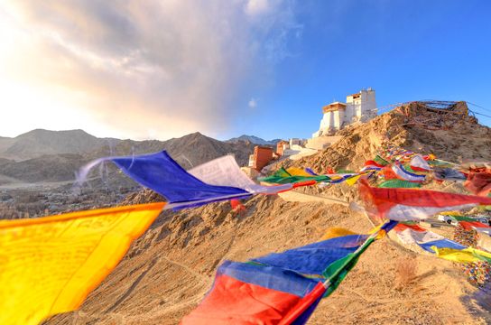 Voyage en Asie — Monastère blanc perché sur une colline aride du Ladakh, entouré de drapeaux de prière colorés flottant au vent au-dessus d’un paysage montagneux désertique.
