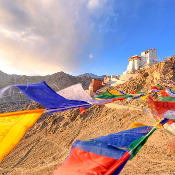 Voyage en Asie — Monastère blanc perché sur une colline aride du Ladakh, entouré de drapeaux de prière colorés flottant au vent au-dessus d’un paysage montagneux désertique.