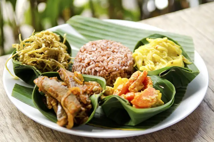 Un plat traditionnel balinais Nasi Campur servi sur une feuille de bananier dans une assiette, composé de riz complet entouré de diverses spécialités locales comme des légumes assaisonnés et des brochettes.