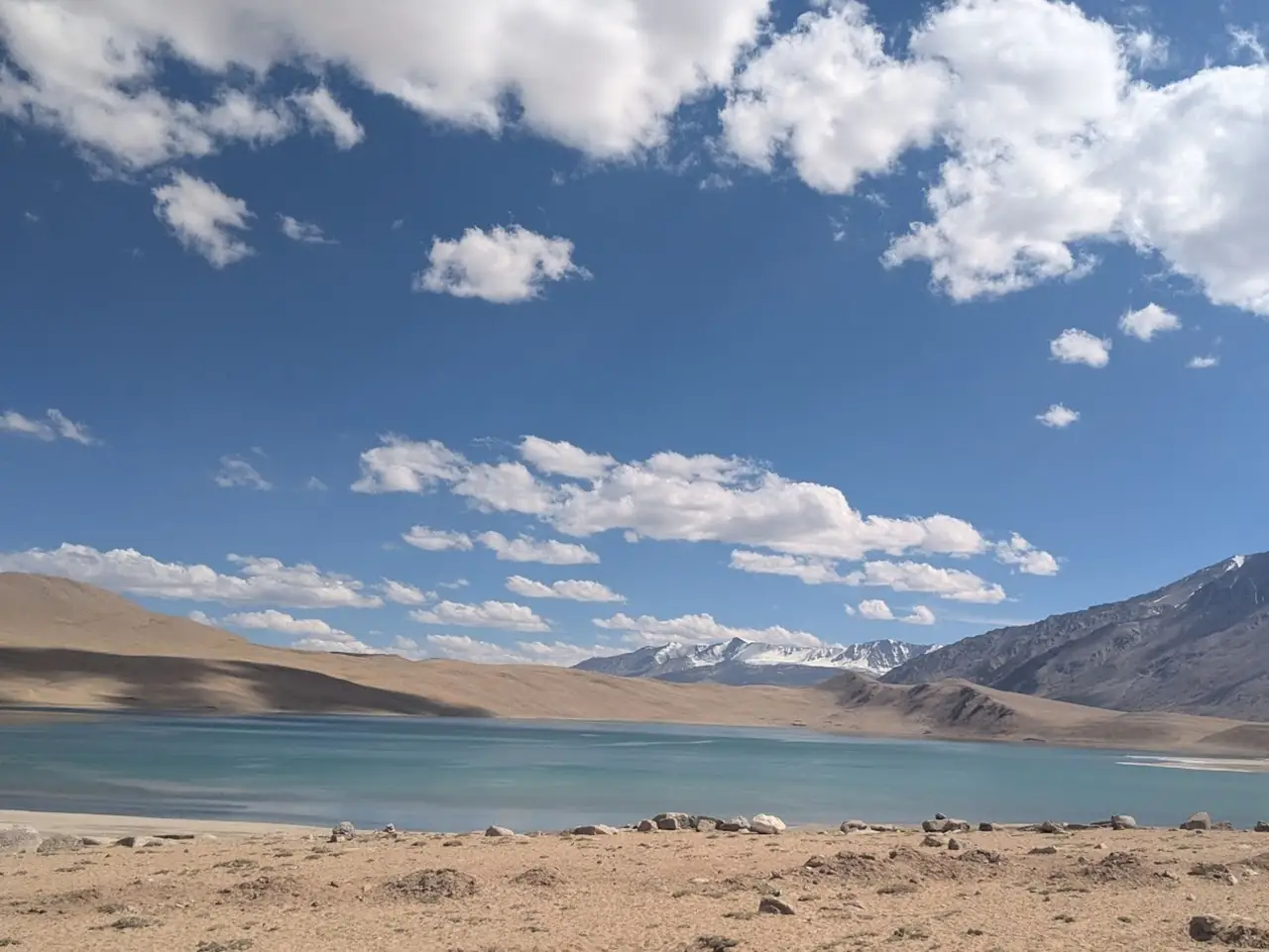 Voyage en Asie — Lac de montagne aux eaux turquoise dans la Shibuk Valley, entouré de collines ocres et dominé par des sommets enneigés sous un ciel bleu lumineux et nuageux.