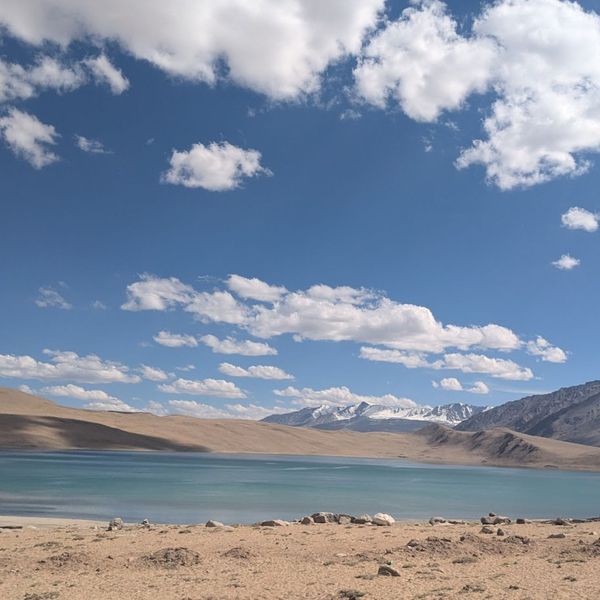 Voyage en Asie — Lac de montagne aux eaux turquoise dans la Shibuk Valley, entouré de collines ocres et dominé par des sommets enneigés sous un ciel bleu lumineux et nuageux.