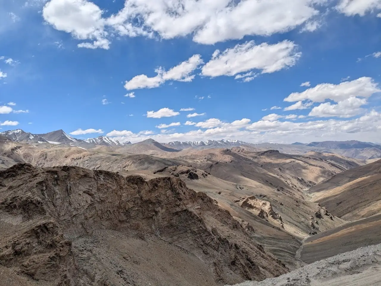 Voyage en Asie — Paysage de montagnes arides et escarpées entre Kyamar et Mandalchan, avec une vallée rocheuse et un ruisseau au premier plan, dominés par des sommets enneigés sous un ciel bleu lumineux.