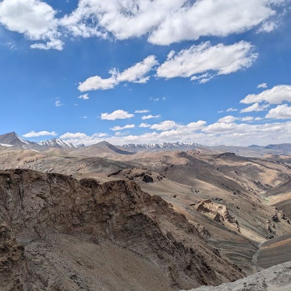 Voyage en Asie — Paysage de montagnes arides et escarpées entre Kyamar et Mandalchan, avec une vallée rocheuse et un ruisseau au premier plan, dominés par des sommets enneigés sous un ciel bleu lumineux.