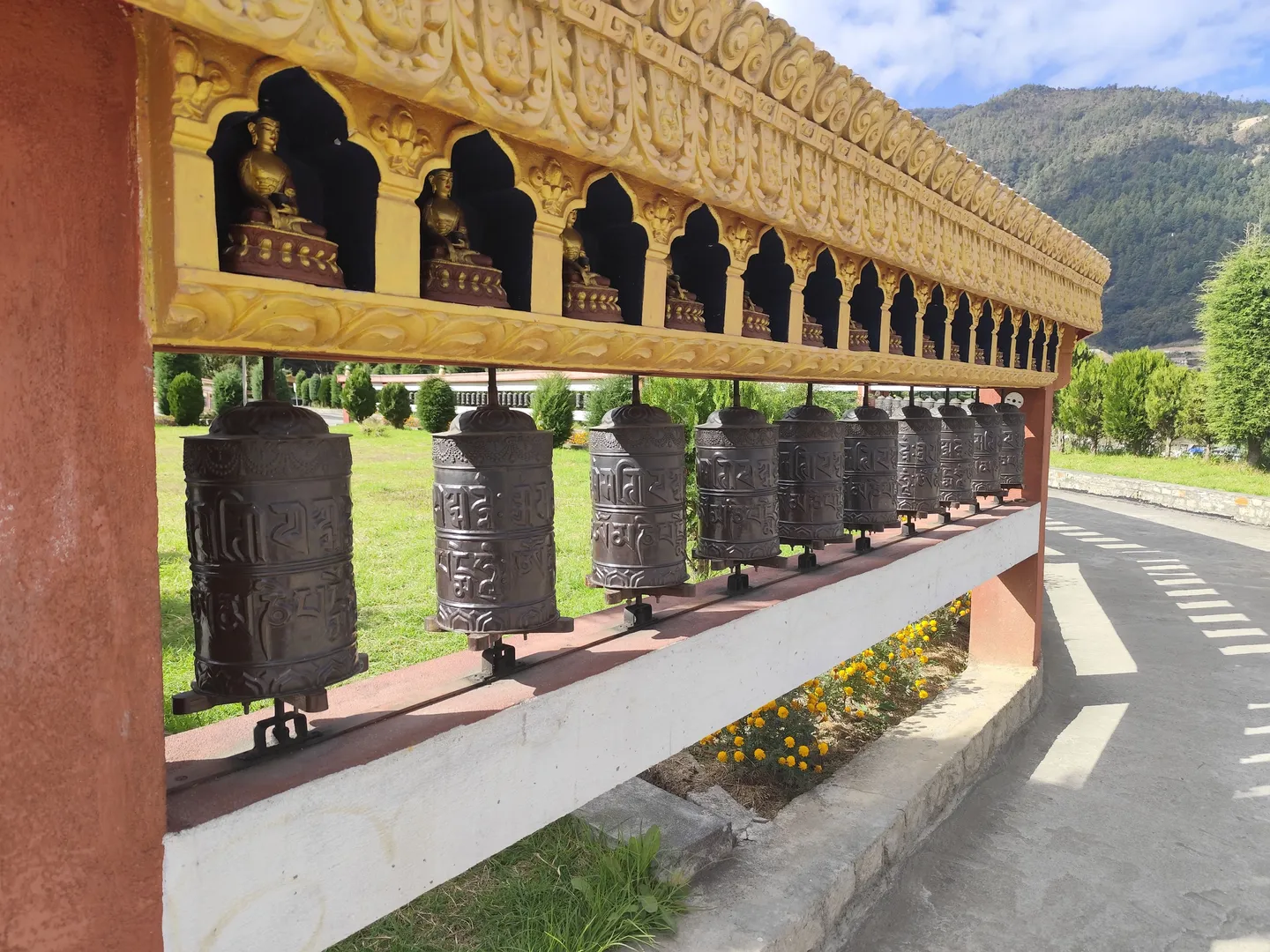 Rangée de moulins à prières tibétains en bronze surmontée d'une structure dorée contenant des statuettes de Bouddha dans des niches, située à Dirang avec un paysage montagneux en arrière-plan
