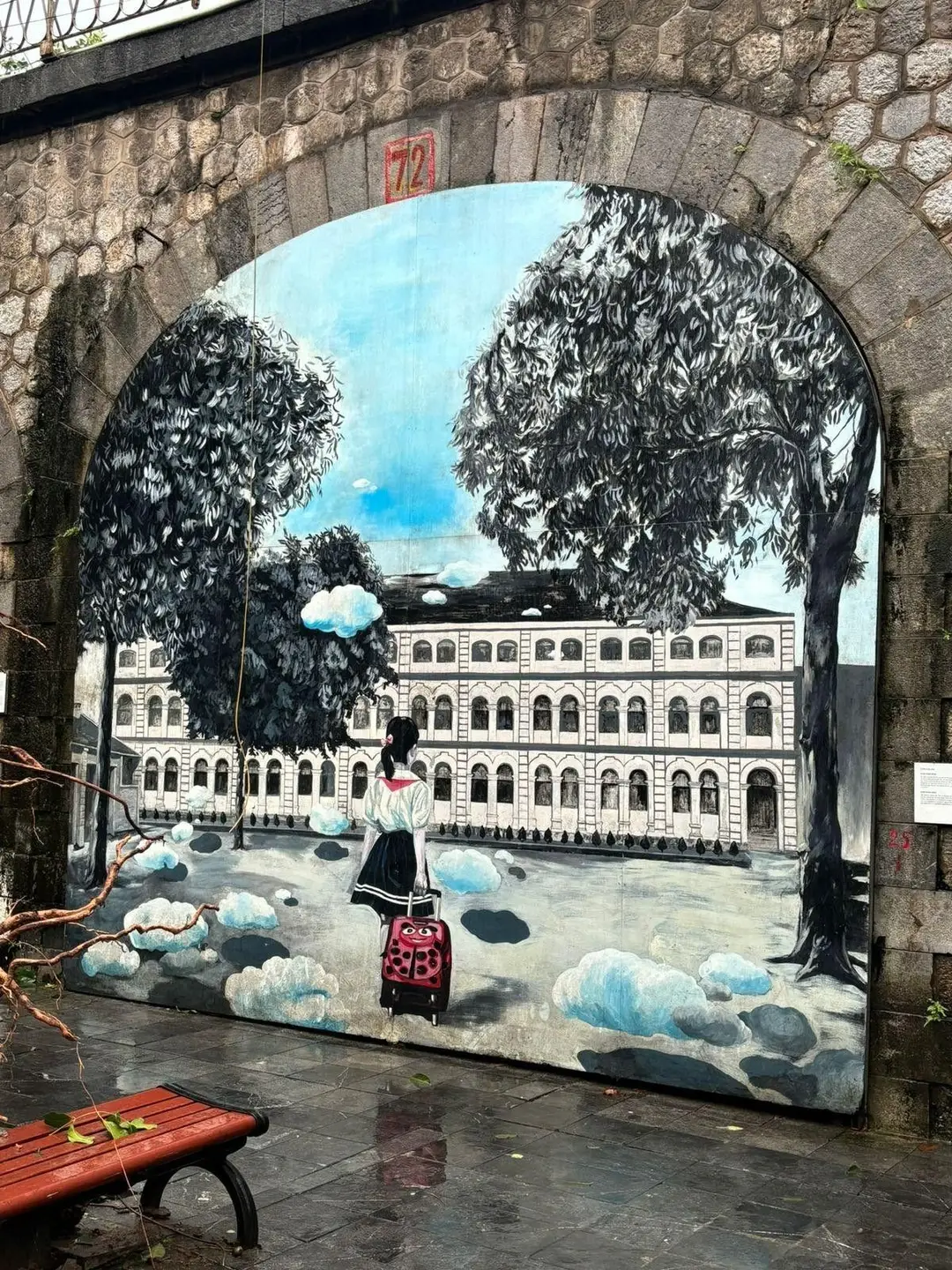 Une peinture murale urbaine sous une arche de la rue Phung Hung à Hanoi, représentant une jeune fille de dos avec une valise rouge face à un grand bâtiment colonial entouré d'arbres.