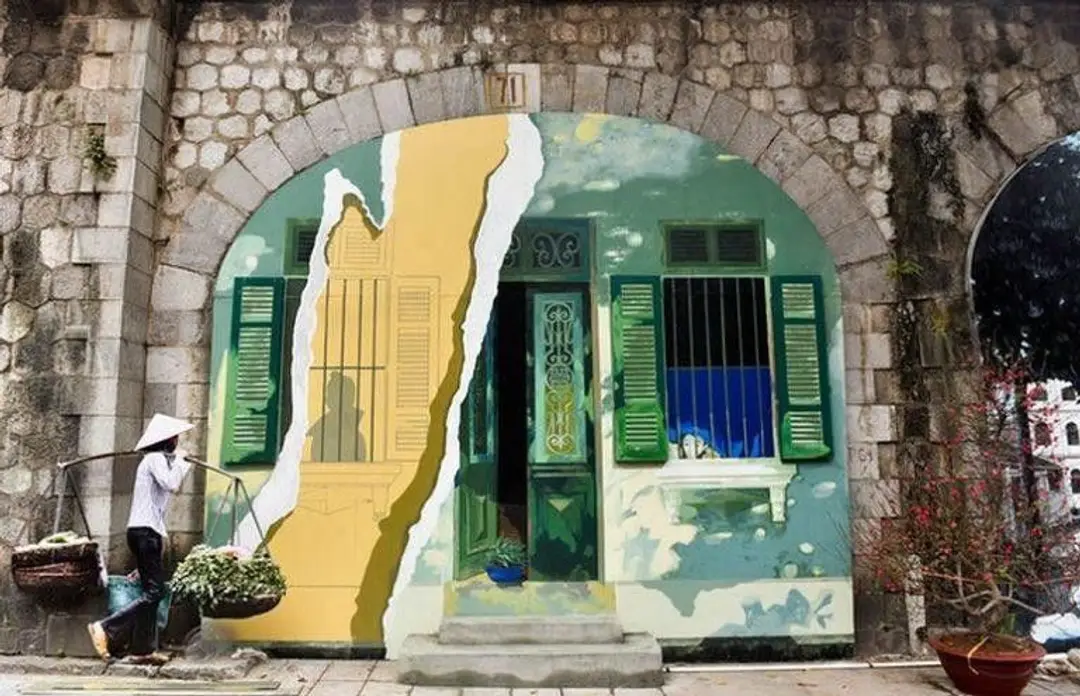 Une fresque murale trompe-l'œil sous l'arche numéro 71 de la rue Phung Hung à Hanoi, représentant une maison traditionnelle aux volets verts entrouverts, devant laquelle passe une vendeuse ambulante portant un chapeau conique et un palanche.