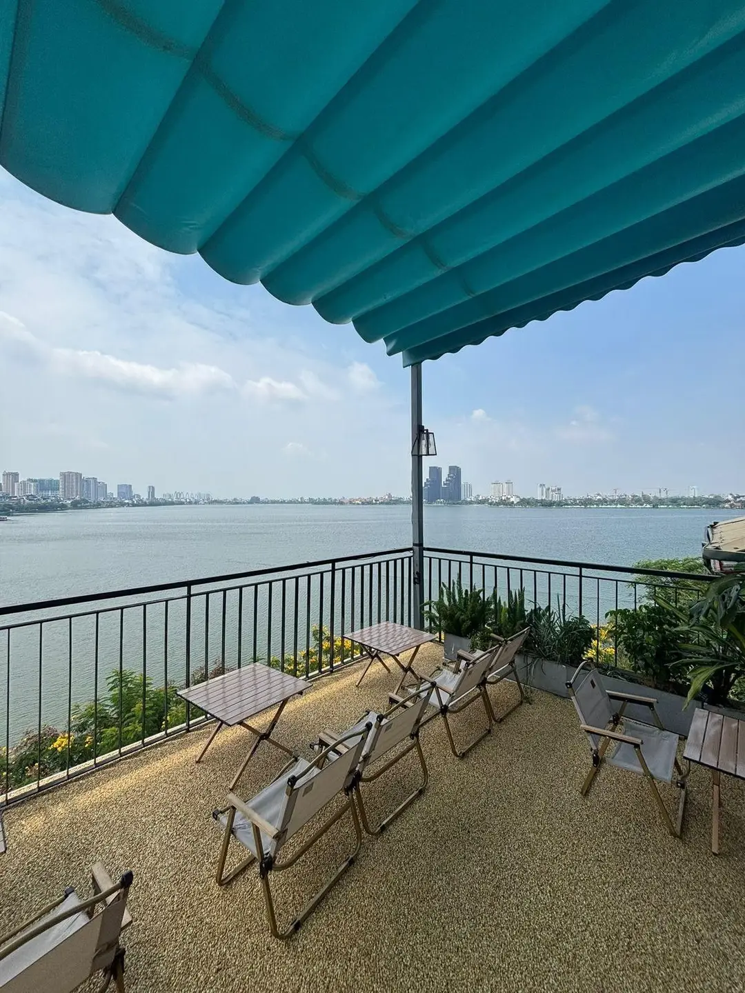 Une terrasse de café en rooftop aménagée avec des chaises et tables pliantes sous un store bleu, offrant une vue panoramique dégagée sur les eaux du lac de l'Ouest et les immeubles de Hanoi à l'horizon.