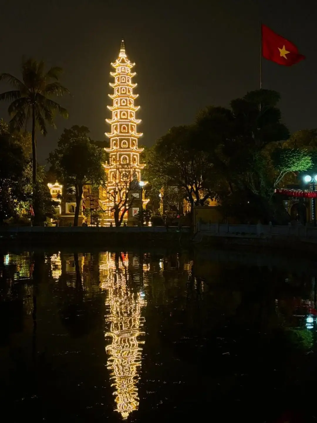 La haute pagode Tran Quoc à plusieurs étages entièrement illuminée de nuit, se reflétant parfaitement sur les eaux sombres du lac de l'Ouest à Hanoi à côté du drapeau vietnamien.