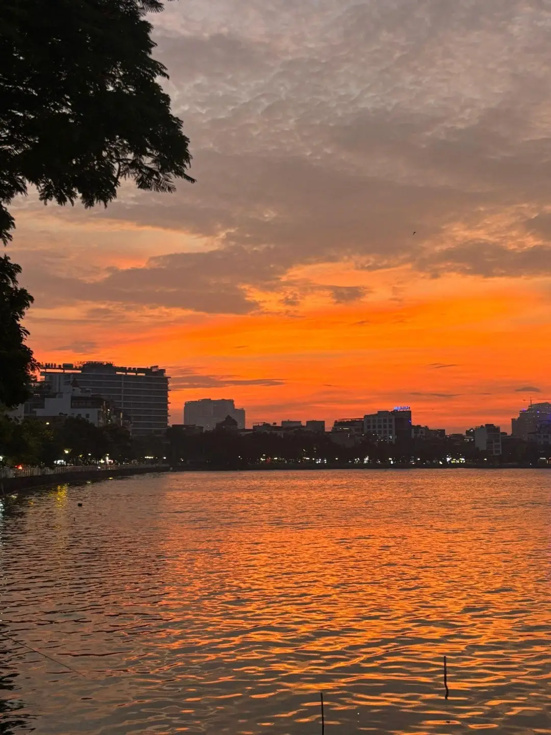 Un ciel flamboyant aux nuances orange et rouge se reflétant sur les eaux calmes du lac de l'Ouest à Hanoi, avec la silhouette de la ville en arrière-plan à la tombée de la nuit.