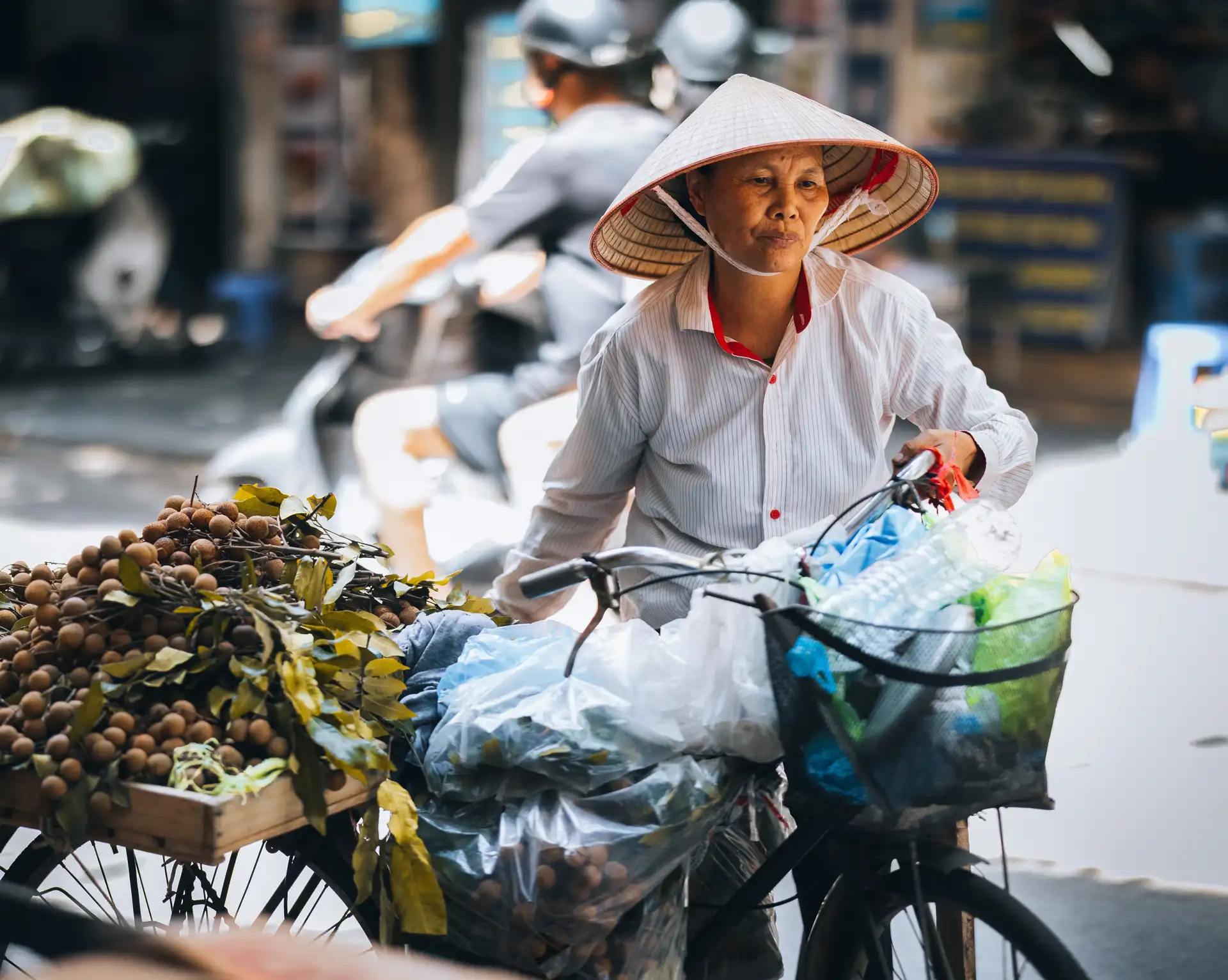une femme vietnamienne portant un chapeau conique traditionnel pousse son velo charge de marchandises dans une rue animee de hanoi