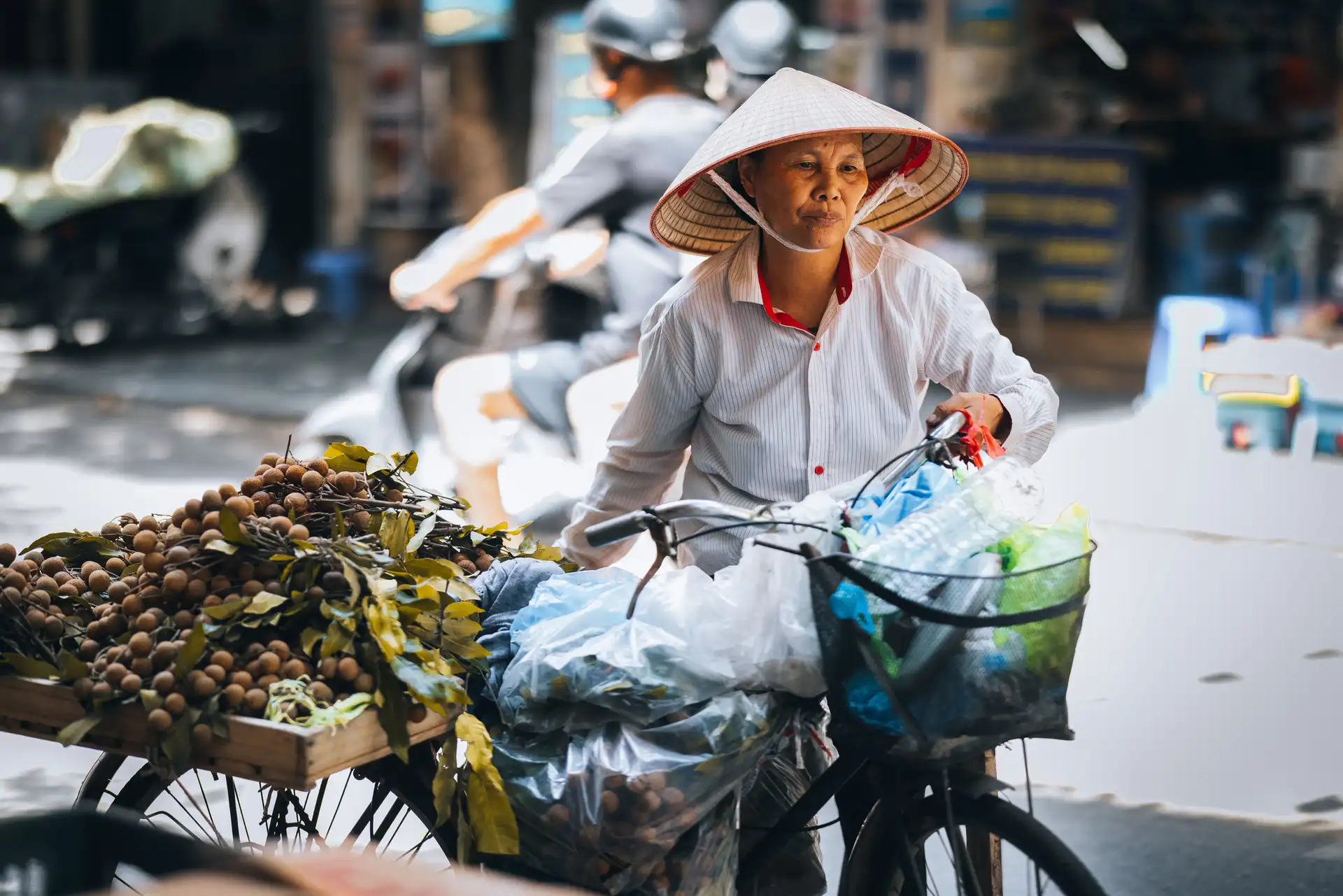 une femme vietnamienne portant un chapeau conique traditionnel pousse son velo charge de marchandises dans une rue animee de hanoi