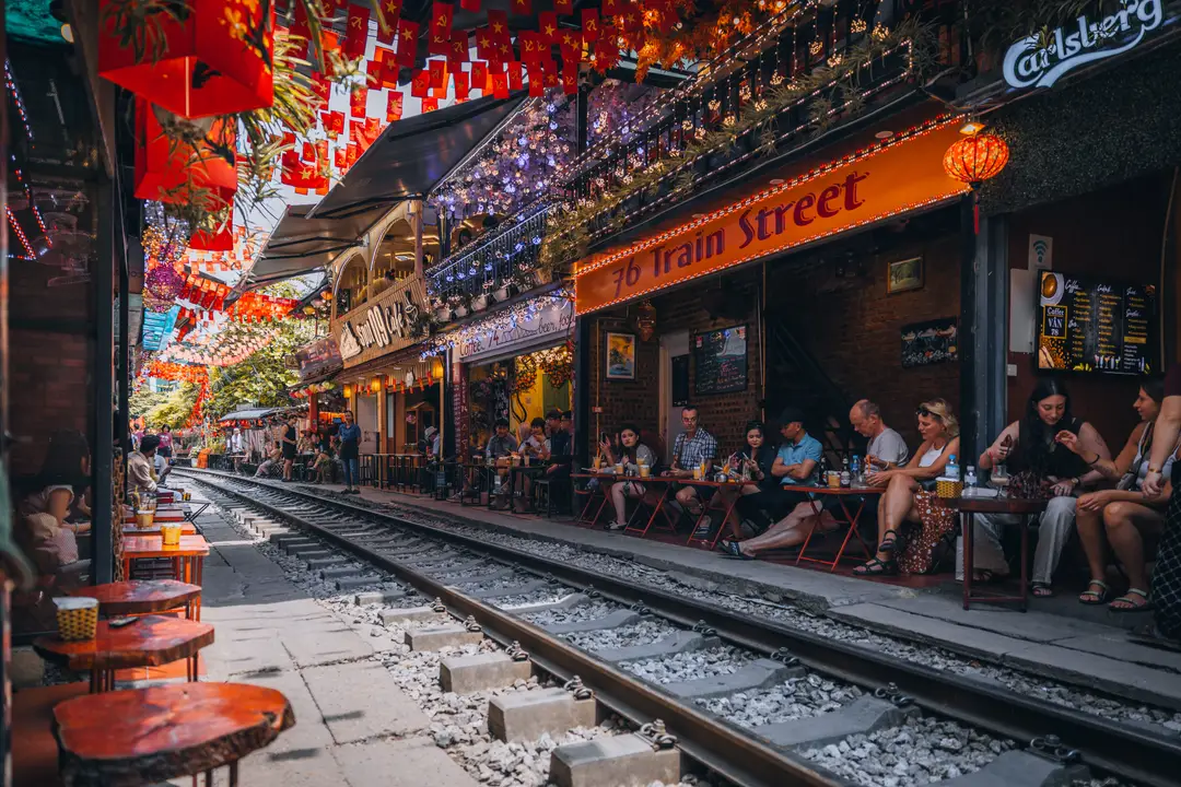 Un train bleu et blanc s'avançant lentement au milieu de la rue étroite de Train Street à Hanoi, alors que les voyageurs assis aux terrasses prennent des photos.