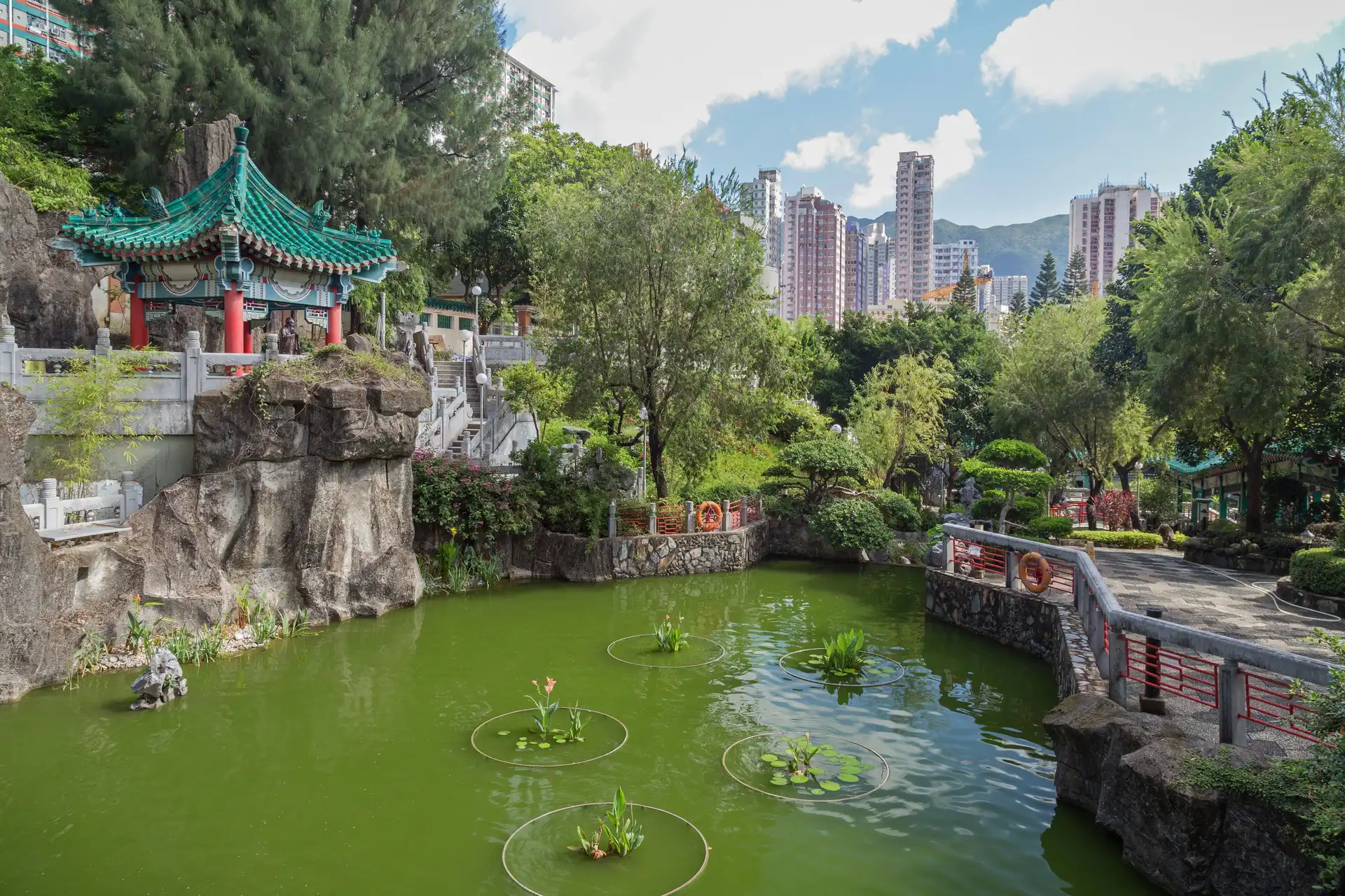 pavillon traditionnel de style chinois avec un toit en tuiles vertes au bord d un etang verdoyant entoure d arbres avec des gratte ciels en arriere plan