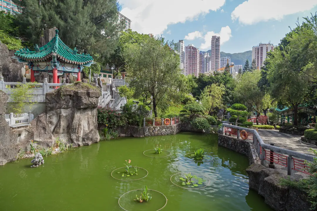 pavillon traditionnel de style chinois avec un toit en tuiles vertes au bord d un etang verdoyant entoure d arbres avec des gratte ciels en arriere plan