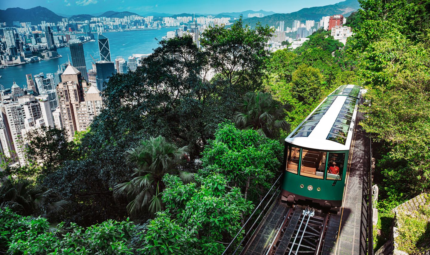 Vue plongeante depuis Victoria Peak montrant le Peak Tram historique de couleur verte montant la colline, entouré d'une végétation luxuriante avec les gratte-ciel de Hong Kong et Victoria Harbour en arrière-plan.