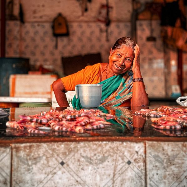 Portrait d'une femme indienne souriante en sari orange et vert assise derrière un étal de crevettes fraîches dans un marché local de Mumbai.