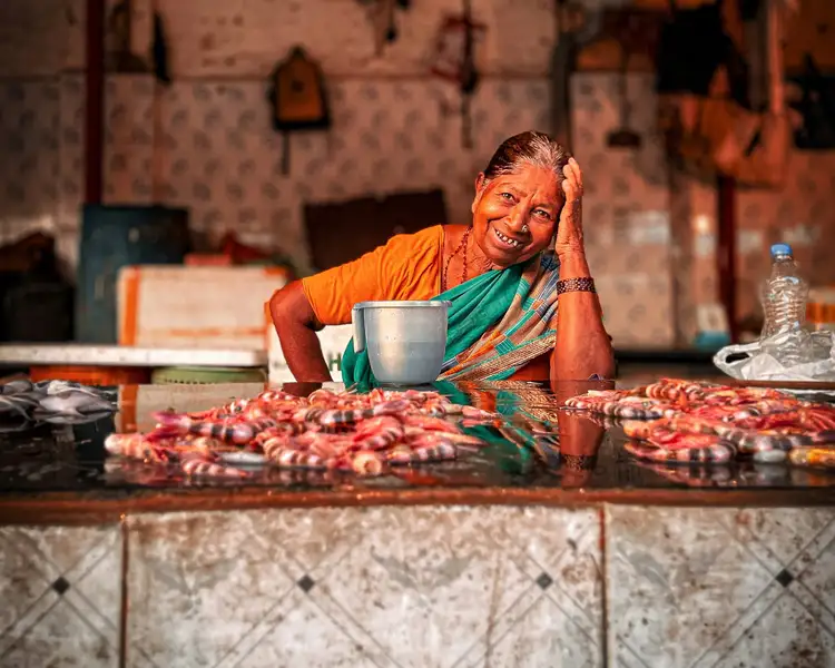Portrait d'une femme indienne souriante en sari orange et vert assise derrière un étal de crevettes fraîches dans un marché local de Mumbai.