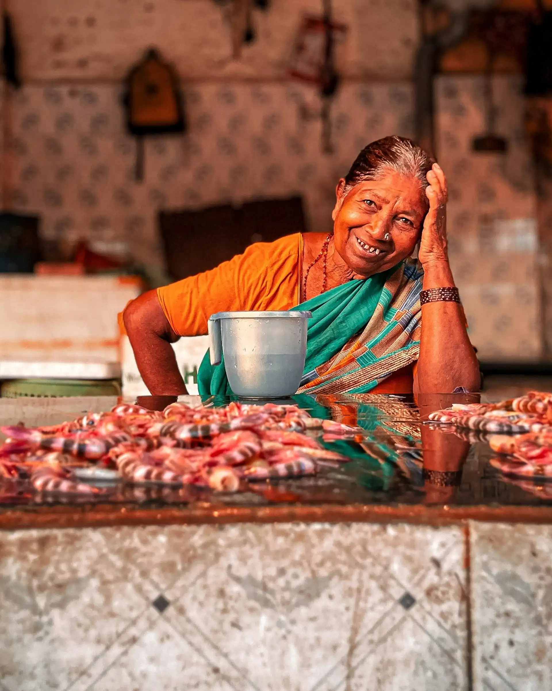 Portrait d'une femme indienne souriante en sari orange et vert assise derrière un étal de crevettes fraîches dans un marché local de Mumbai.