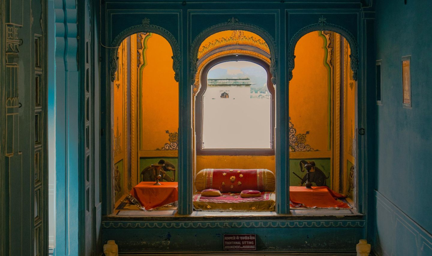 Vue d'une alcôve royale traditionnelle dans un palais à Udaipur avec des arches sculptées bleues, des murs jaunes ornés de peintures et des coussins rouges confortables.
