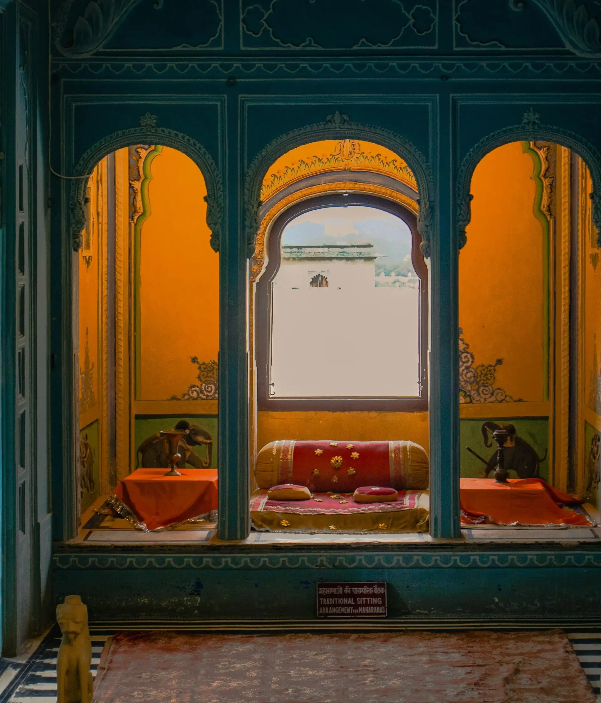 Vue d'une alcôve royale traditionnelle dans un palais à Udaipur avec des arches sculptées bleues, des murs jaunes ornés de peintures et des coussins rouges confortables.