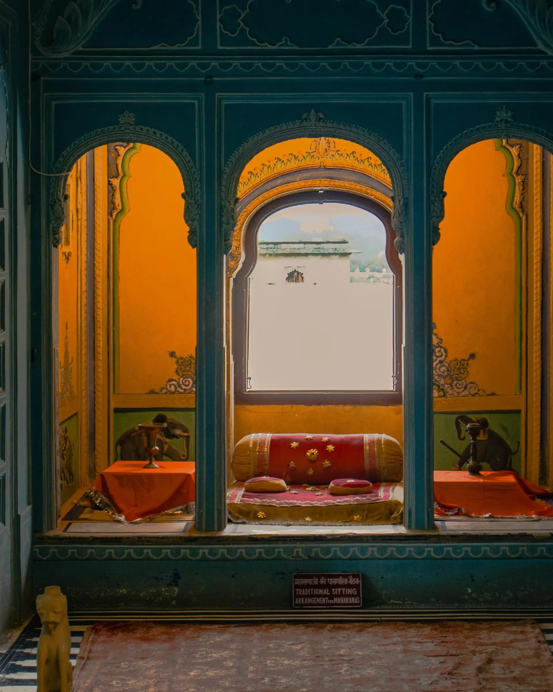Vue d'une alcôve royale traditionnelle dans un palais à Udaipur avec des arches sculptées bleues, des murs jaunes ornés de peintures et des coussins rouges confortables.