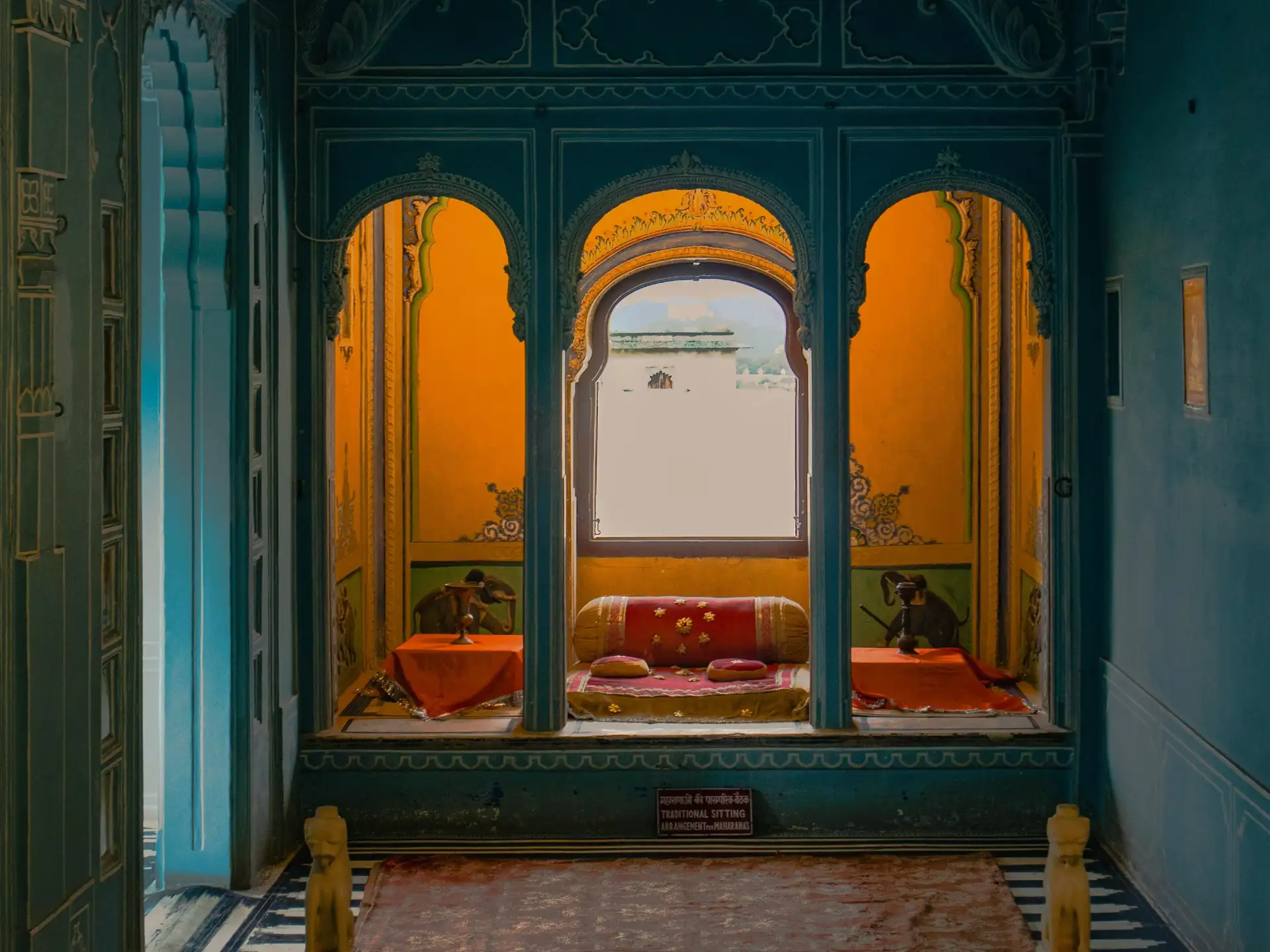 Vue d'une alcôve royale traditionnelle dans un palais à Udaipur avec des arches sculptées bleues, des murs jaunes ornés de peintures et des coussins rouges confortables.
