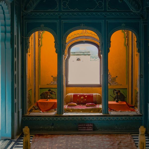 Vue d'une alcôve royale traditionnelle dans un palais à Udaipur avec des arches sculptées bleues, des murs jaunes ornés de peintures et des coussins rouges confortables.