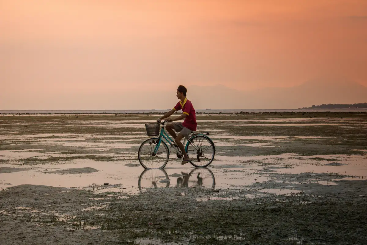 Silhouette d'un cycliste pédalant sur le sable mouillé d'une plage à marée basse sous un ciel teinté de rose et d'orange au coucher du soleil à Bali.