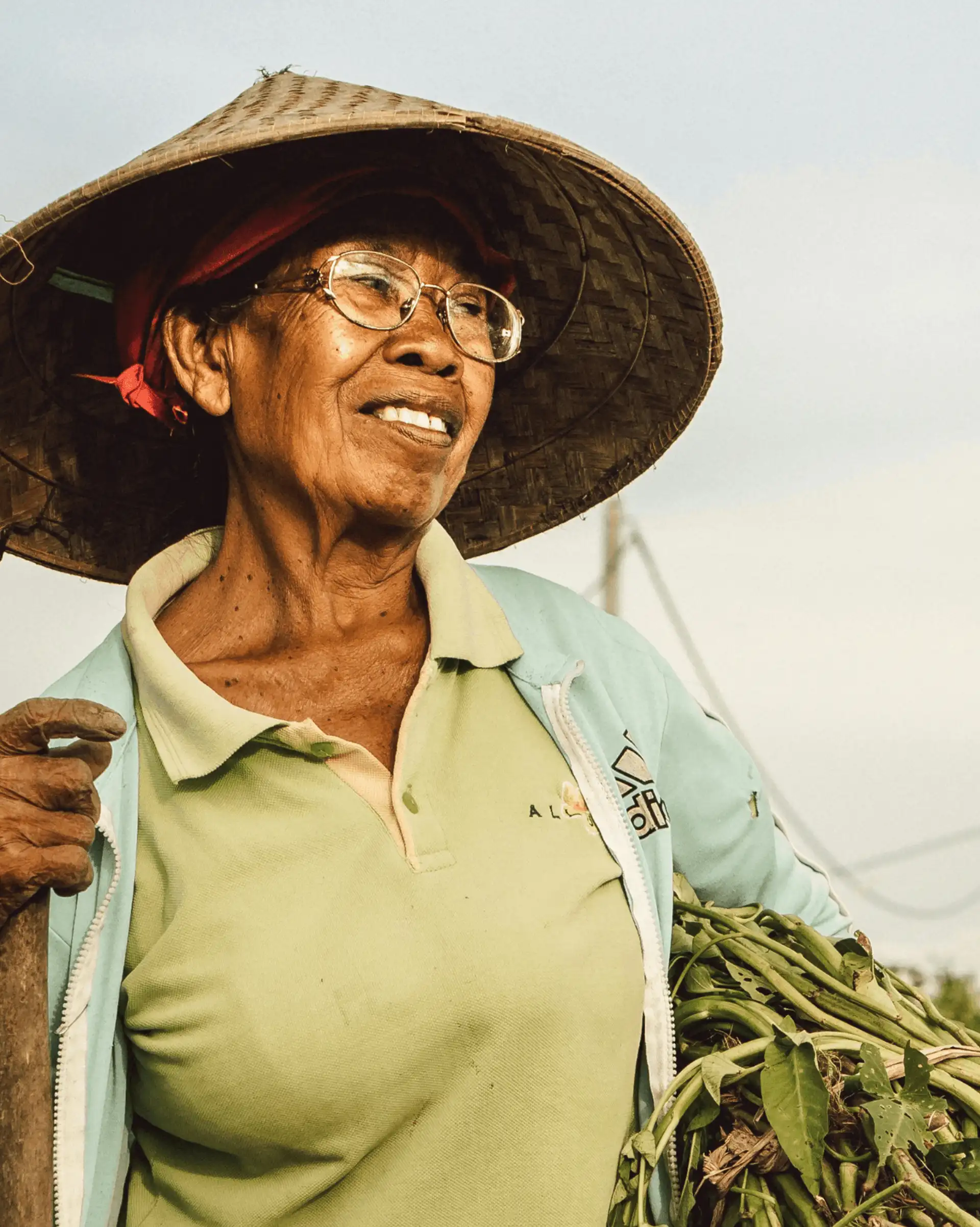 Voyage en Asie — Portrait d'une femme indonésienne âgée portant un chapeau conique traditionnel, souriante et en plein travail agricole à Bali, dans un cadre rural authentique.
