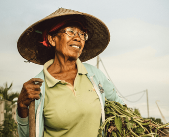 Voyage en Asie — Portrait d'une femme indonésienne âgée portant un chapeau conique traditionnel, souriante et en plein travail agricole à Bali, dans un cadre rural authentique.