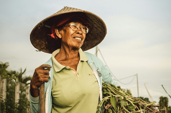 Voyage en Asie — Portrait d'une femme indonésienne âgée portant un chapeau conique traditionnel, souriante et en plein travail agricole à Bali, dans un cadre rural authentique.